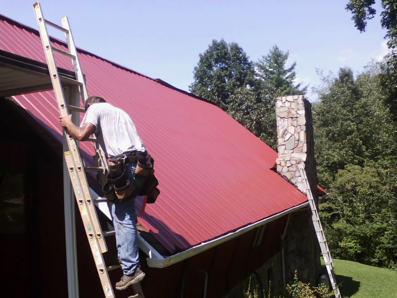 Roofer on a ladder beside a red metal roof and stone chimney — Soffit & Fascia Repair by Roofing Brothers in Alsip
